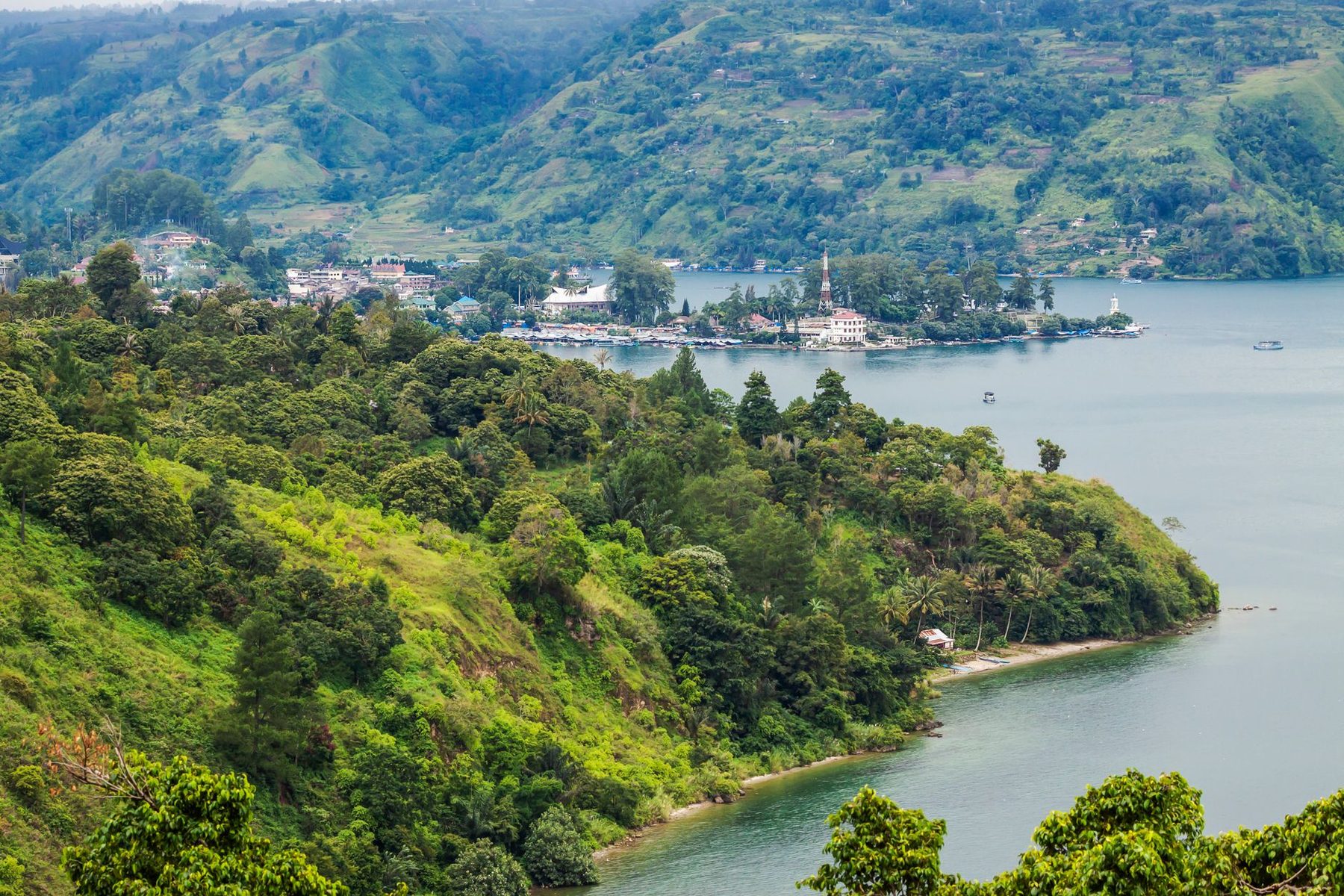 Bukit hijau rimbun menghadap danau tenang dengan desa di kejauhan